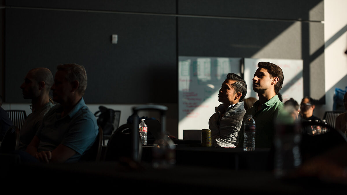 People sit in a dimly lit room, attentively listening to a presentation by Denver Headshot Co. There are water bottles and notepads on the table in front of them.